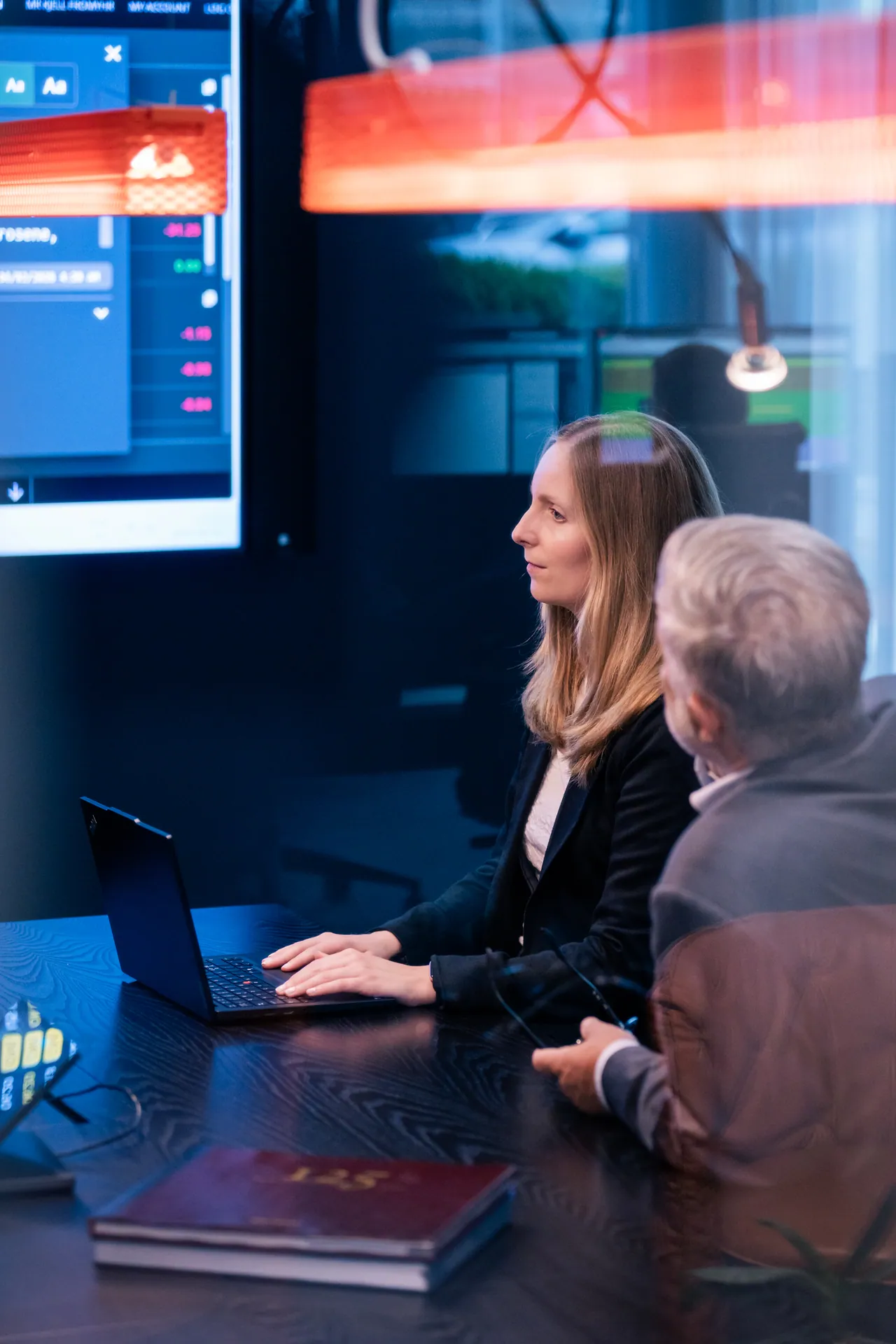 Two colleagues at a meeting table, with market screens and warm red light in the background.